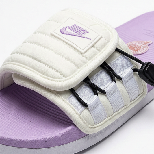 Nike slide crema morada vista detallada angular close-up 3/4 frontal-izquierda correa puffer quilted cage bandas blancas horizontales tabs negros cordón negro doble stopper parche Nike swoosh morado superior-izquierda footbed morado lavanda emblema rojo dorado derecho